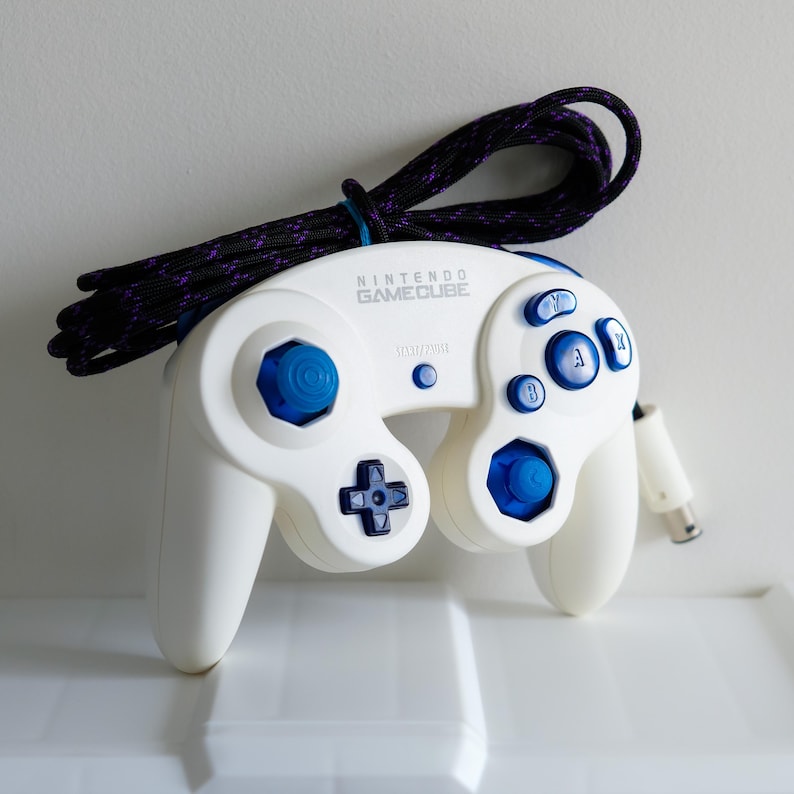 May include: White Nintendo GameCube controller with blue buttons and a black and purple braided cable. The controller has the text "NINTENDO GAMECUBE" printed on the top.