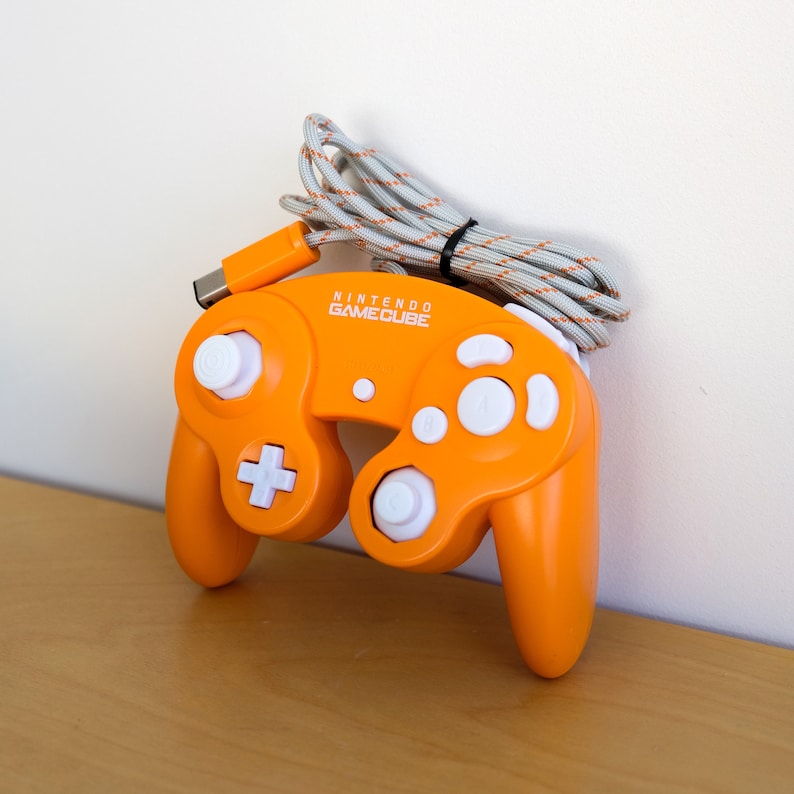 May include: An orange Nintendo GameCube controller with white buttons and a white cord. The controller has the words "Nintendo GameCube" printed on the front.