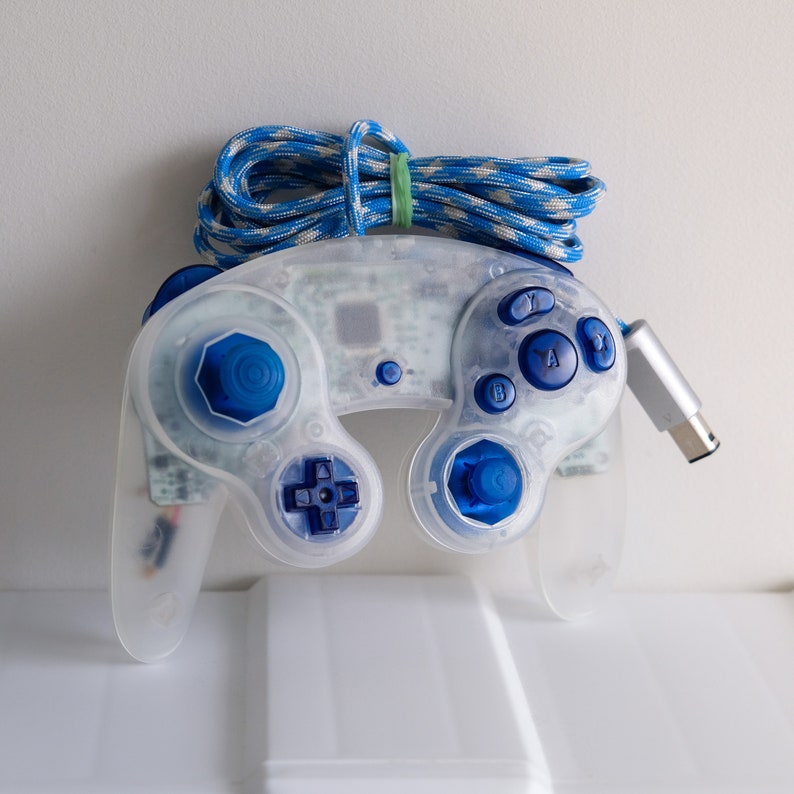 May include: A clear white GameCube controller with blue buttons. The controller has a blue and white striped cord attached to it.