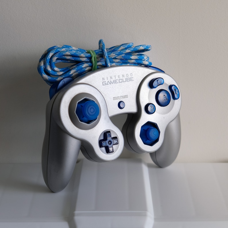 May include: A silver GameCube controller with blue buttons and a blue and white braided cord. The controller has the text "NINTENDO GAMECUBE" printed on the top.