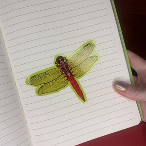 K&ouml;nnte beinhalten: Ein gelber und roter Libellenaufkleber mit detaillierten Fl&uuml;geln und einem roten K&ouml;rper. Der Aufkleber befindet sich auf einer linierten Notizbuchseite.
