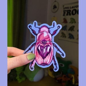 May include: A pink and purple beetle sticker with a heart-shaped design. The sticker is on a light blue background.