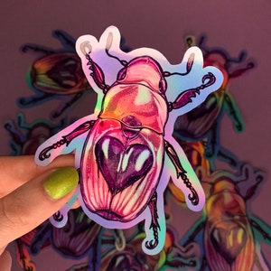 Puede incluir: Una pegatina holográfica de un escarabajo rosa y morado con un diseño en forma de corazón en la espalda. La pegatina es iridiscente y tiene un acabado brillante.