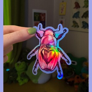 May include: A holographic sticker of a pink and iridescent beetle with a heart-shaped design on its back.