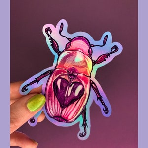 May include: A holographic sticker of a pink and purple beetle with a heart-shaped design on its back.