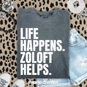 May include: A dark gray t-shirt with the text "LIFE HAPPENS. ZOLOFT HELPS." in white block letters. The shirt is folded and placed on a leopard print surface, with a pair of denim shorts and white sneakers visible.