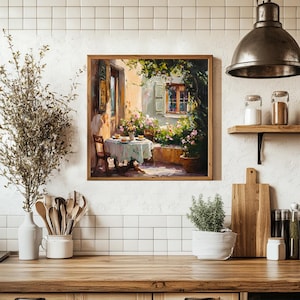 May include: A painting depicting a patio scene with a table set for a meal, surrounded by lush greenery and a stone building with a window. The table is covered with a white tablecloth and has a vase of flowers in the centre. The painting is framed in a wooden frame.