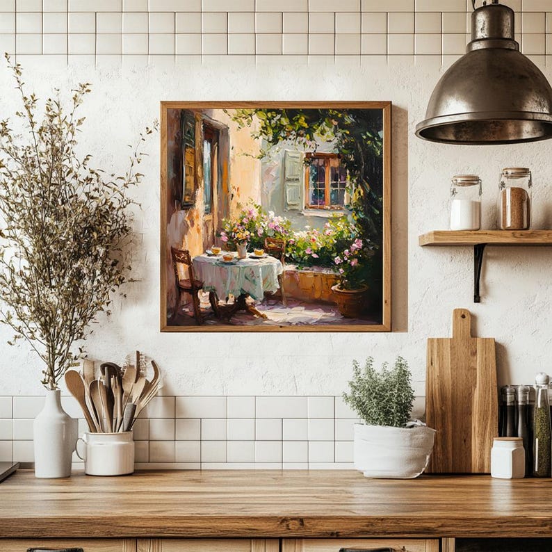 May include: A painting depicting a patio scene with a table set for a meal, surrounded by lush greenery and a stone building with a window. The table is covered with a white tablecloth and has a vase of flowers in the centre. The painting is framed in a wooden frame.