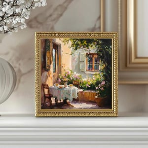 May include: A framed oil painting depicting a quaint outdoor dining scene. A table with a white tablecloth is set with plates and cups, surrounded by lush greenery and a charming stone building with a window. The painting is framed in a gold-coloured frame.