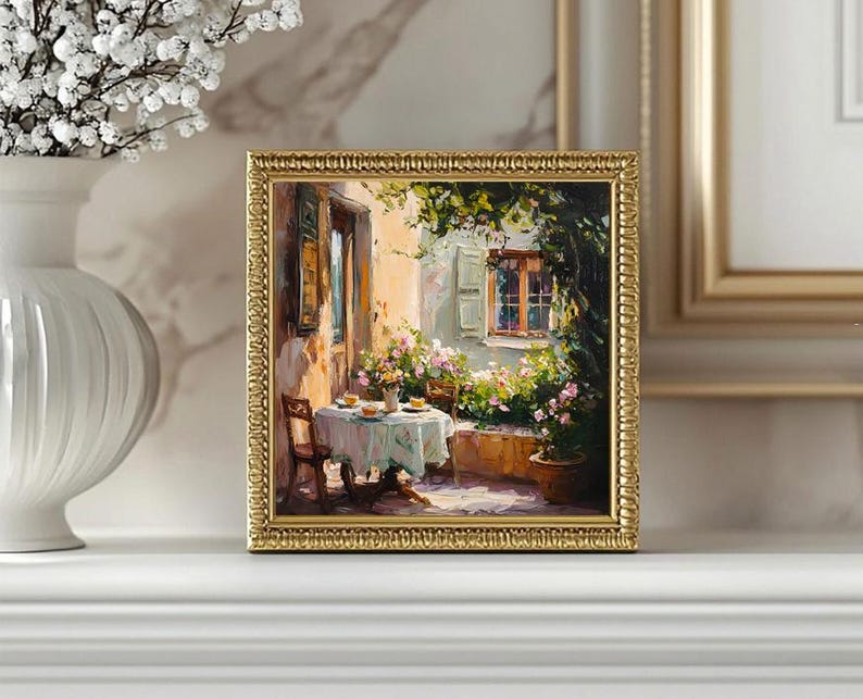 May include: A framed oil painting depicting a quaint outdoor dining scene. A table with a white tablecloth is set with plates and cups, surrounded by lush greenery and a charming stone building with a window. The painting is framed in a gold-coloured frame.