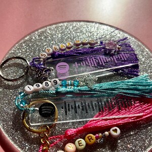 May include: Three clear acrylic keychains with colourful beaded tassels. Each keychain features a Spotify logo and a barcode-like design. The top keychain has purple beads and the text "SPEAK NOW". The middle keychain has turquoise beads and the numbers "1989". The bottom keychain has pink beads and the text "LOVER".