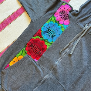 Peut inclure: Un sweat à capuche gris avec un motif floral brodé coloré sur le devant. Les fleurs sont dans des tons de rose, bleu, rouge, vert et jaune.