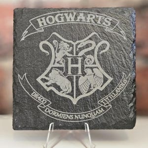 May include: Square, dark grey slate coaster with the Hogwarts crest etched into the surface. The crest features a lion, eagle, badger, and snake surrounding the letter "H", with the words "Hogwarts", "Draco Dormiens Nunquam Titillandus" in a banner.