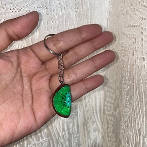 Puede incluir: Un llavero con un pequeño dije semicircular en forma de rodaja de kiwi. El dije tiene un interior verde brillante con pequeñas semillas negras y una corteza marrón. Está unido a una argolla de metal plateado mediante una cadena corta.