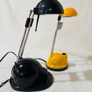 May include: Two adjustable desk lamps, one black and one yellow, each featuring a dome-shaped head, a chrome-finished telescoping arm, and a rounded base with a power switch. The black lamp is positioned in the foreground, while the yellow lamp sits slightly behind it, both connected by black power cords.