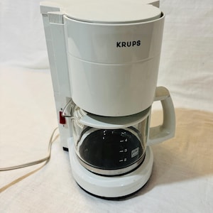 May include: White Krups coffee maker with a glass carafe. The carafe has measurements for 2, 3, and 4 cups of coffee. The coffee maker has a handle and a red on/off switch. The brand name "KRUPS" is printed on the front.