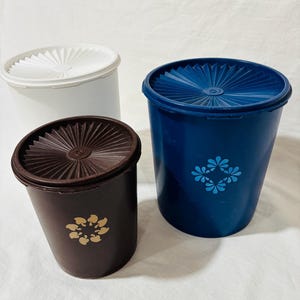 May include: Three vintage plastic containers with lids. One is white, one is dark brown, and one is blue. The brown and blue containers have a floral design. The lids have a radial design.