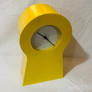 May include: A bright yellow, modern-style desk clock. The clock has a circular face with black hands and a white background. The clock's body is a solid, geometric shape with a rounded top and a rectangular base.