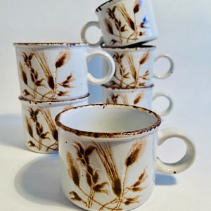 May include: A stack of six white ceramic mugs with a brown wheat design. The mugs have a brown rim and a brown handle.