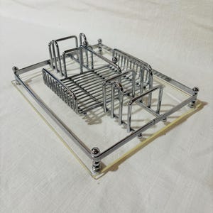 May include: A chrome-plated metal and clear acrylic vanity tray. The tray has a rectangular shape with rounded corners and small ball feet. The metal portion features a soap dish and toothbrush holder.