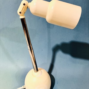 May include: Cream-colored desk lamp with a cylindrical head and a spherical base. The lamp features a chrome-colored adjustable arm and a power cord. The lamp is designed for focused lighting and has a retro aesthetic.