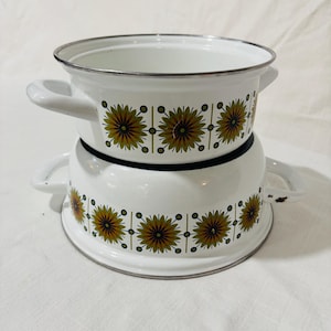 May include: Two white enamel pots with handles, stacked. Each pot features a repeating pattern of yellow and brown stylized flowers with green accents. The rims and handles are white, and the edges are silver.