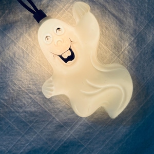 May include: A white, translucent, ghost-shaped light cover attached to a black electrical cord. The ghost figure features a printed face with two eyes, a nose, and an open mouth showing two prominent front teeth, illuminated from within by a light.