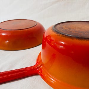 May include: Two orange-red enameled cast iron saucepans. One saucepan has a long handle. The pans have a gradient color effect, transitioning from a darker red at the base to a brighter orange at the top. The interior is a darker brown.