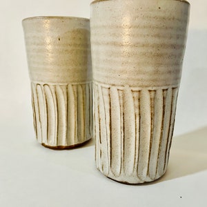 May include: Two white ceramic mugs with a textured, ribbed design. The mugs are slightly off-white with a subtle brown glaze.