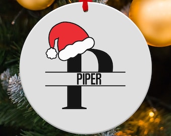 May include: White round Christmas ornament with a black letter "P" with a red and white Santa hat on top. The name "PIPER" is written in black letters on a white banner below the letter.
