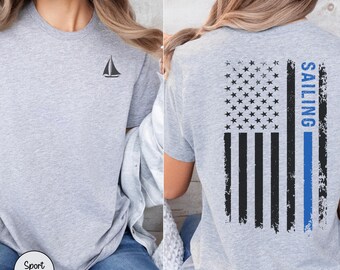 Może przedstawiać: Koszulka w kolorze sport grey z małą czarną grafiką żaglówki na lewej piersi. Z tyłu znajduje się postarzany wzór flagi amerykańskiej w kolorze czarnym z pionowym niebieskim paskiem i napisem SAILING wydrukowanym na niebiesko.