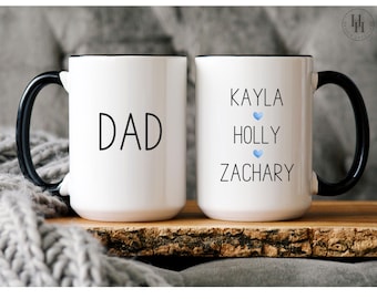 Może przedstawiać: Dwa białe ceramiczne kubki z czarnymi uchwytami i czarnymi obwódkami. Kubek po lewej stronie ma napis "DAD" czarnym tekstem. Kubek po prawej stronie ma napis "KAYLA HOLLY ZACHARY" czarnym tekstem z niebieskim symbolem serca po każdym imieniu.