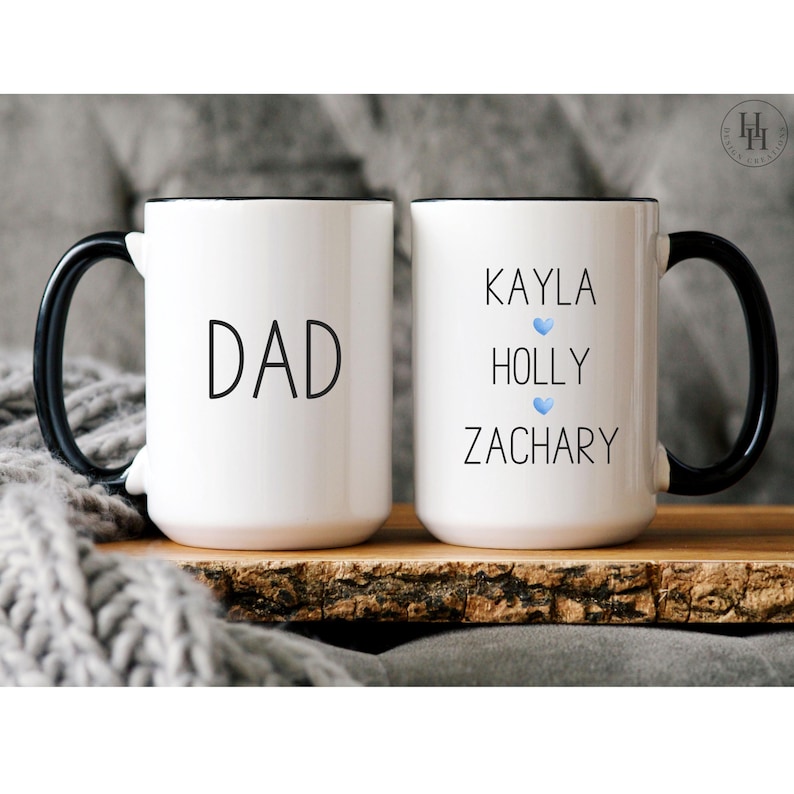 Może przedstawiać: Dwa białe ceramiczne kubki z czarnymi uchwytami i czarnymi obw&oacute;dkami. Kubek po lewej stronie ma napis "DAD" czarnym tekstem. Kubek po prawej stronie ma napis "KAYLA HOLLY ZACHARY" czarnym tekstem z niebieskim symbolem serca po każdym imieniu.