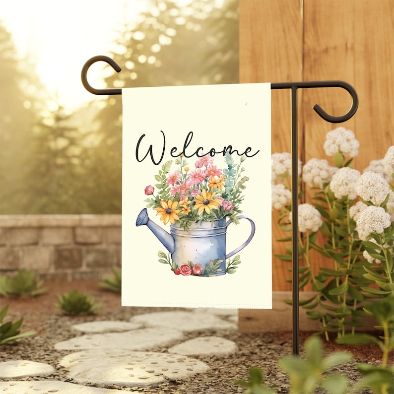May include: A welcome garden flag with a white background and a blue watering can filled with colorful flowers. The text "Welcome" is written in black cursive lettering.