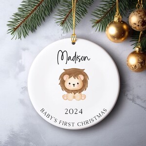 May include: White ceramic Christmas ornament with a brown lion illustration, the text "Madison", "Baby's First Christmas", and the year "2024".