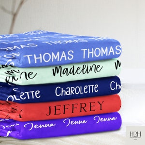 May include: A stack of five personalized fleece blankets in various colors. The top blanket is blue with the name "Thomas" repeated in white. The second blanket is light blue with the name "Madeline" in black. The third blanket is dark blue with the name "Charlotte" in white. The fourth blanket is red with the name "Jeffrey" in black. The bottom blanket is purple with the name "Jenna" repeated in white.