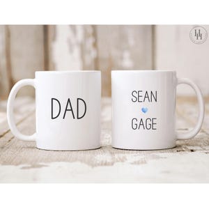 Może przedstawiać: Dwa białe ceramiczne kubki z czarnym tekstem. Jeden kubek ma napis "DAD", a drugi kubek ma napis "SEAN GAGE" z niebieskim sercem.