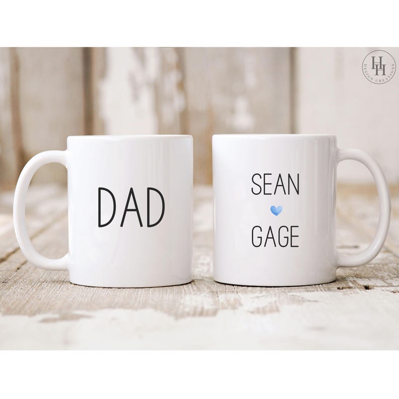 Może przedstawiać: Dwa białe ceramiczne kubki z czarnym tekstem. Jeden kubek ma napis "DAD", a drugi kubek ma napis "SEAN GAGE" z niebieskim sercem.