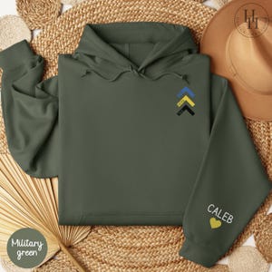 May include: A military green hoodie with a yellow, black, and blue arrow design on the front and the word "Caleb" with a yellow heart on the left sleeve.