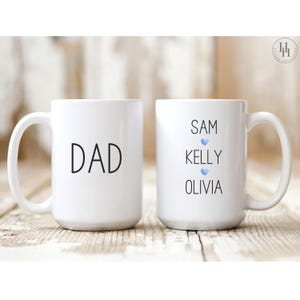 Może przedstawiać: Dwa białe ceramiczne kubki z czarnym tekstem. Jeden kubek ma napis "DAD", a drugi kubek ma napis "SAM KELLY OLIVIA" z niebieskim sercem między każdym imieniem.