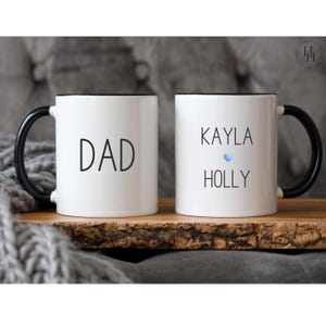 Może przedstawiać: Dwa białe ceramiczne kubki z czarnymi uchwytami i czarnymi obw&oacute;dkami. Jeden kubek ma napis "DAD", a drugi kubek ma napis "KAYLA HOLLY" z niebieskim sercem.
