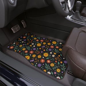 May include: Black car floor mat with a colorful floral pattern. The design features yellow, orange, purple, and white flowers with green leaves on a black background. The mat is in a car interior.