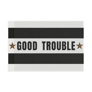Puede incluir: Un objeto decorativo rectangular con un patrón de rayas blancas y negras. La frase "GOOD TROUBLE" está impresa en letras negras en negrita en el centro, flanqueada por estrellas marrones. El objeto tiene una estética limpia y moderna.