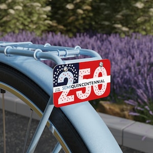 Può includere: Bicicletta azzurra con targa decorativa rossa e blu. La targa presenta il numero "250" in bianco, la scritta "SEMIQUINCENTENNIAL" e gli anni "1776 2026". Fiori viola sullo sfondo.