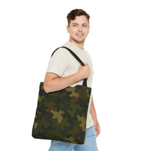 Puede incluir: Un bolso tote con un estampado de camuflaje en tonos verdes, marrones y beige. Tiene una correa negra y se lleva al hombro. El bolso está hecho de un material resistente.