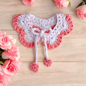 May include: A white and pink crocheted collar with a scalloped edge and a bow tie. The collar has two long ties that end in pom-poms.