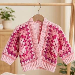 Peut inclure: Cardigan rose en crochet, avec un motif de carrés granny dans différentes nuances de rose. Le vêtement à devant ouvert a un ourlet et des poignets côtelés. Il est présenté sur un cintre en bois. Fait main.