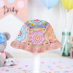Könnte beinhalten: Ein mehrfarbiger Häkelhut mit floralem Muster. Der Hut ist in Pastelltönen wie Rosa, Blau, Gelb und Grün gehalten. Er wird auf einem weißen Ständer präsentiert, wahrscheinlich ein Hut für ein Baby oder Kind, ideal für einen besonderen Anlass.