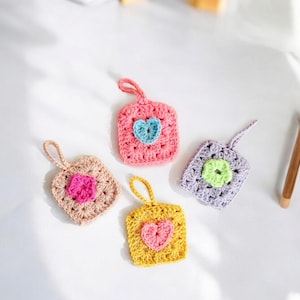 May include: Four crocheted square ornaments with heart designs. The ornaments are in pink, yellow, purple, and beige with different colored hearts.