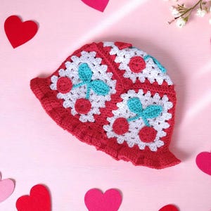 Peut inclure: Un chapeau au crochet rouge et blanc avec un motif de cerises. Le chapeau présente un bord festonné et est orné de fleurs blanches, de cerises rouges et de feuilles turquoise. C'est un article fait à la main.